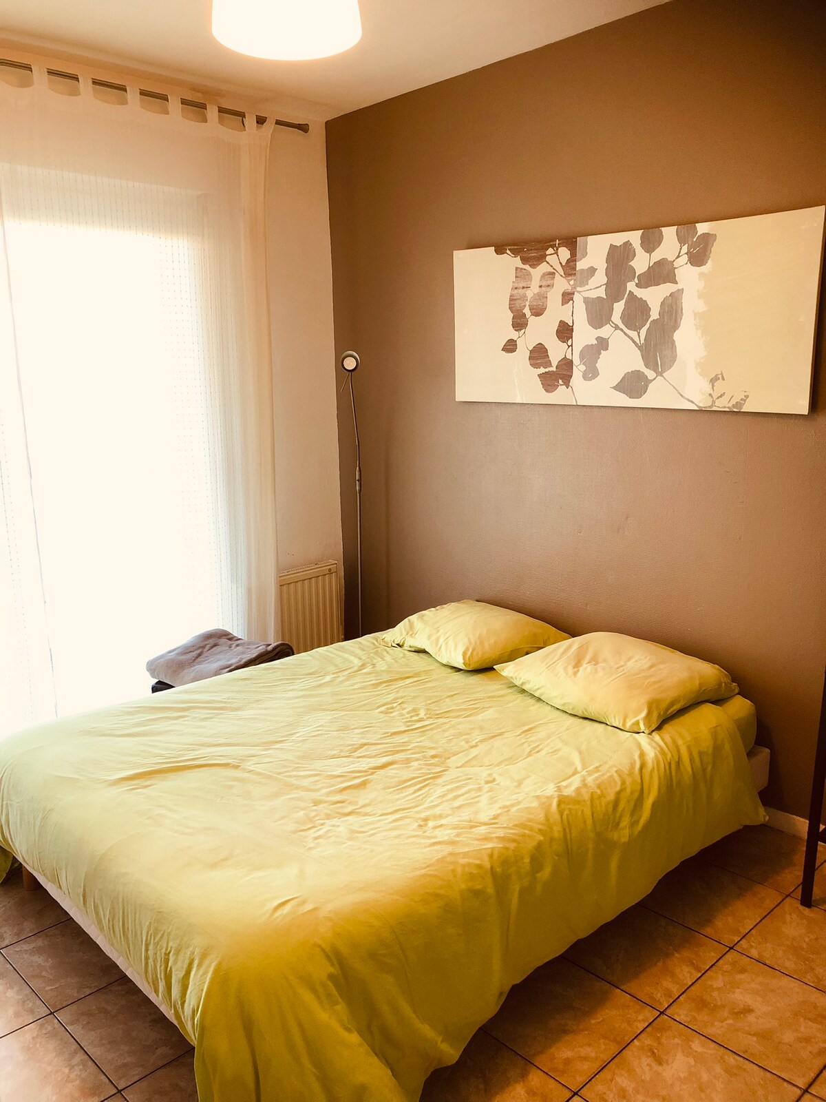 A spacious sleeping area features a light green bedding set laid over a large bed. A piece of wall art with botanical motifs is displayed above the bed. Natural light enters through a sliding glass door, complemented by a floor lamp beside the bed.