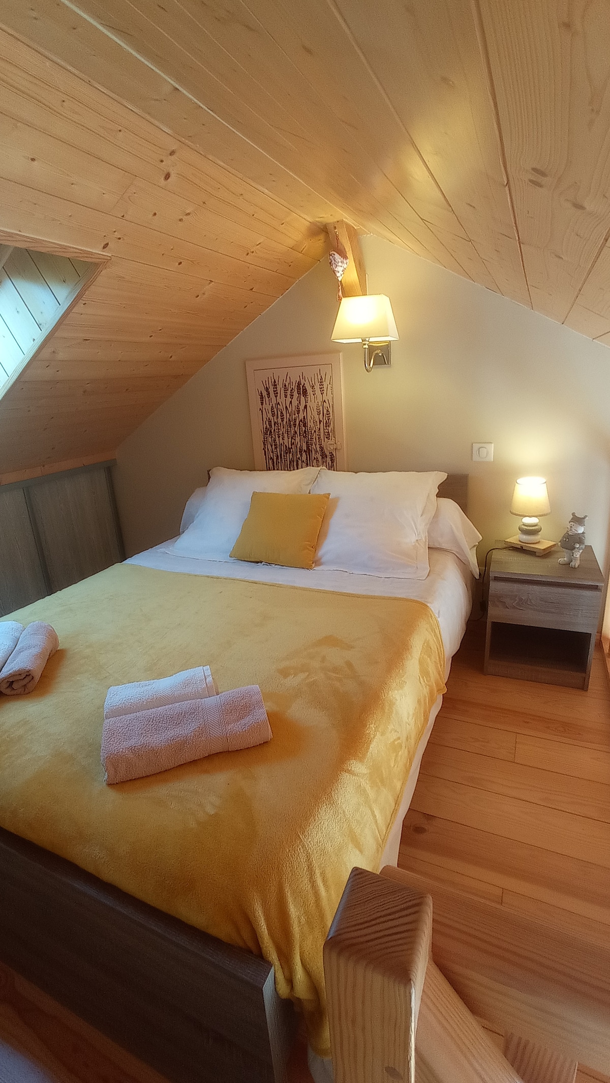 A cozy sleeping area features a spacious bed dressed in crisp white linens with a soft yellow blanket. A pair of rolled towels is neatly arranged on the bed. Soft lighting from wall sconces creates a warm ambiance against the wood-paneled walls.