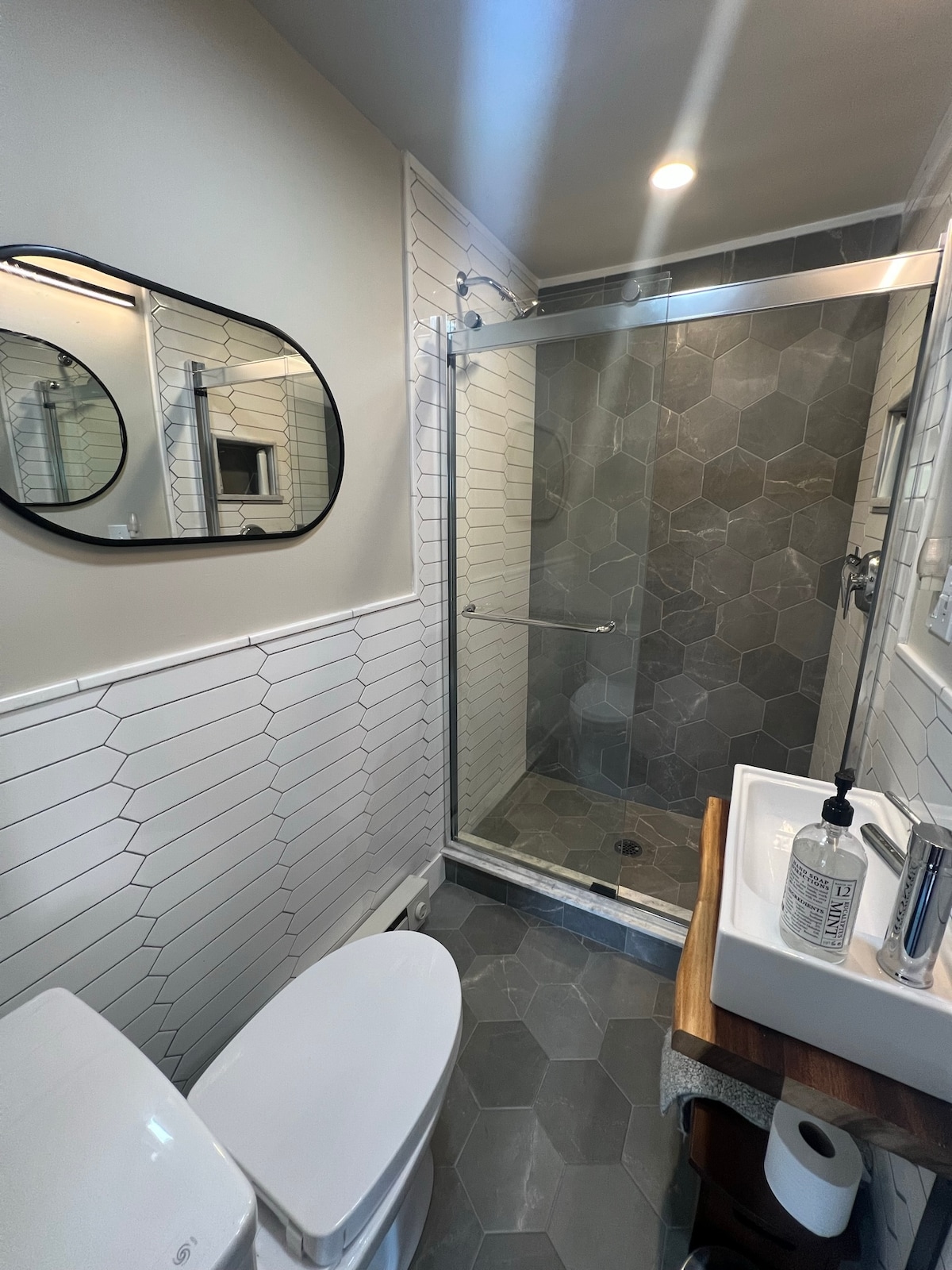 A contemporary bathroom features a sleek glass shower with a hexagonal tiled floor. A modern toilet is positioned beside a stylish sink with a minimalist wooden shelf underneath. Natural light is provided through a frosted window accented by a round mirror.