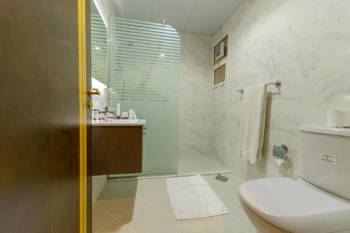 A modern bathroom features elegant marble walls and a glass shower enclosure. A wooden vanity with sink is equipped with toiletries and a large mirror. A white towel is neatly arranged on the floor, and the space is well-lit.