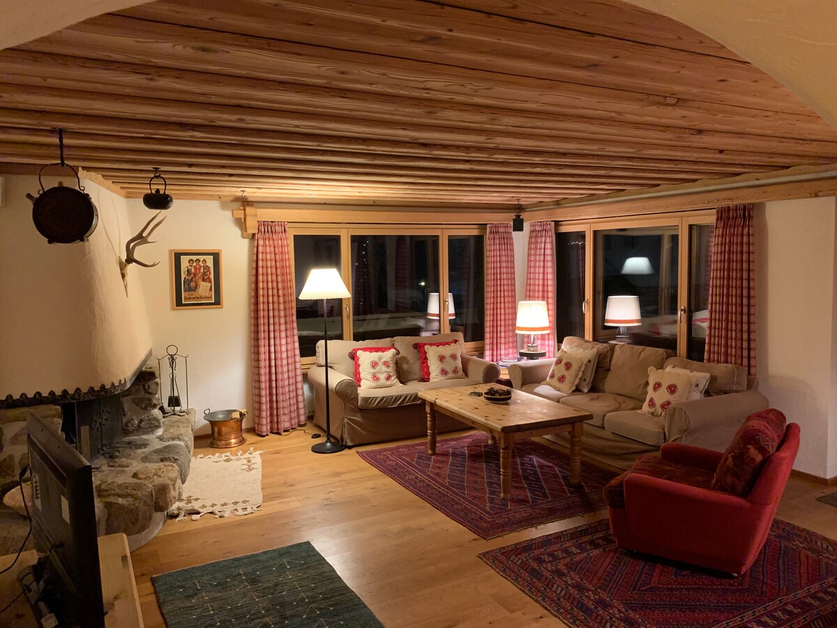 A spacious living area features wooden beams on the ceiling and large windows with checkered curtains. Comfortable seating is arranged around a coffee table, with a stone fireplace providing a cozy focal point. Warm lighting enhances the inviting atmosphere.