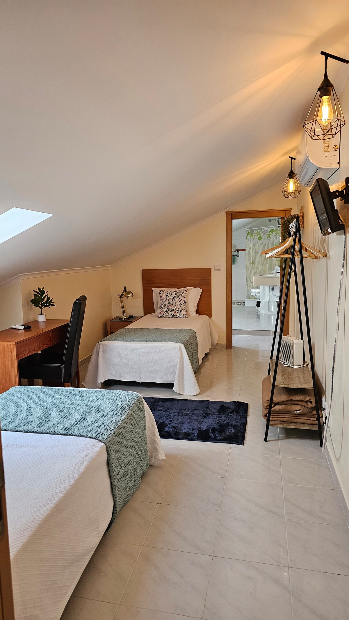 A bright room features two single beds with crisp bedding and a decorative pillow. A desk is positioned beside a window, while a small clothing rack and a cozy rug add functionality. Soft lighting is provided by overhead fixtures, creating a comfortable environment.