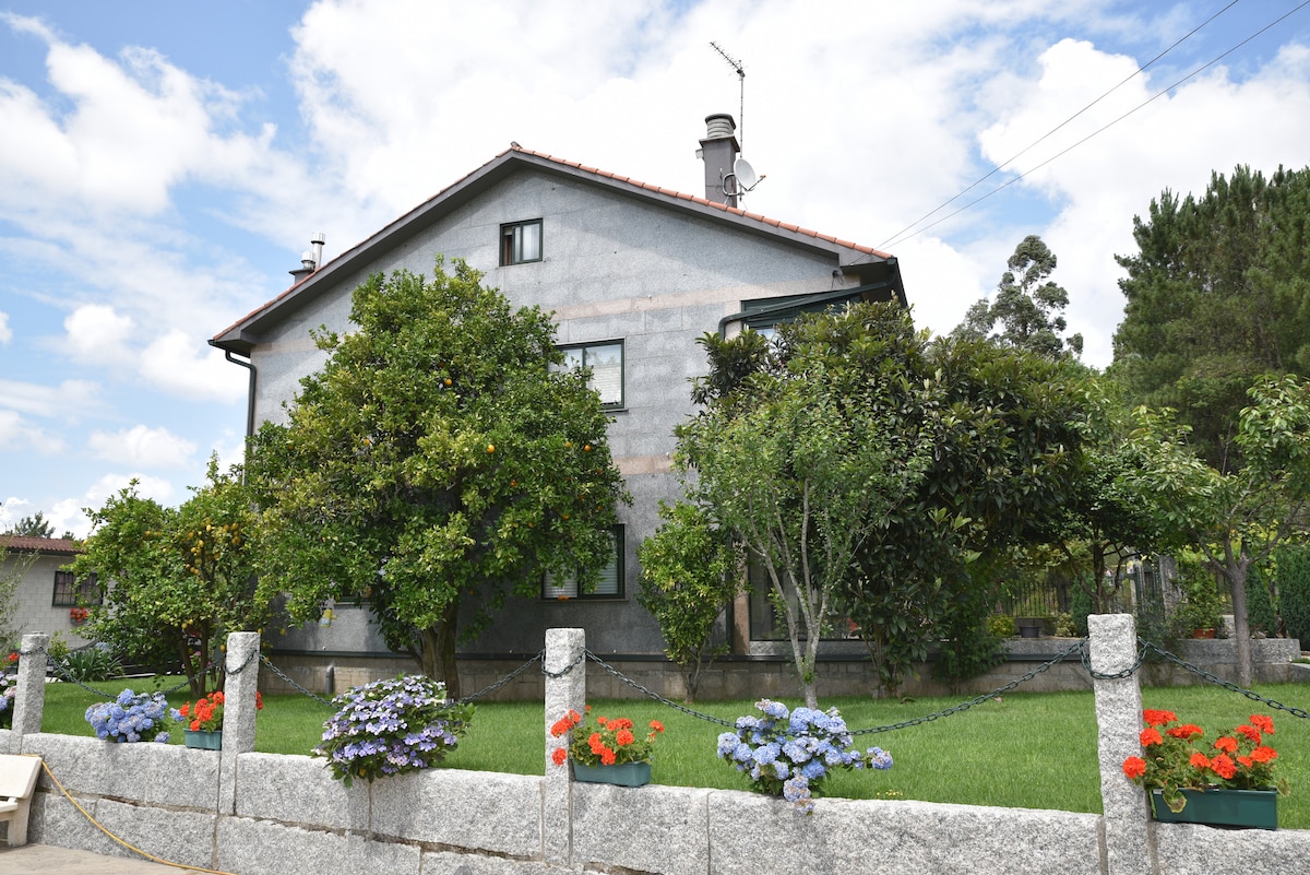 A two-story building is surrounded by greenery, featuring trees and blooming flowers along a stone fence. The yard is spacious with manicured grass, and the home is characterized by a textured exterior and a sloped roof.