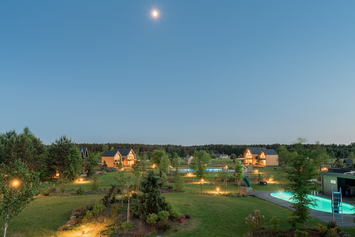 A serene landscape showcases multiple homes illuminated at twilight, surrounded by lush greenery and trees. Two swimming pools glisten under the night sky, while soft lighting highlights pathways and garden areas, creating a tranquil ambiance.