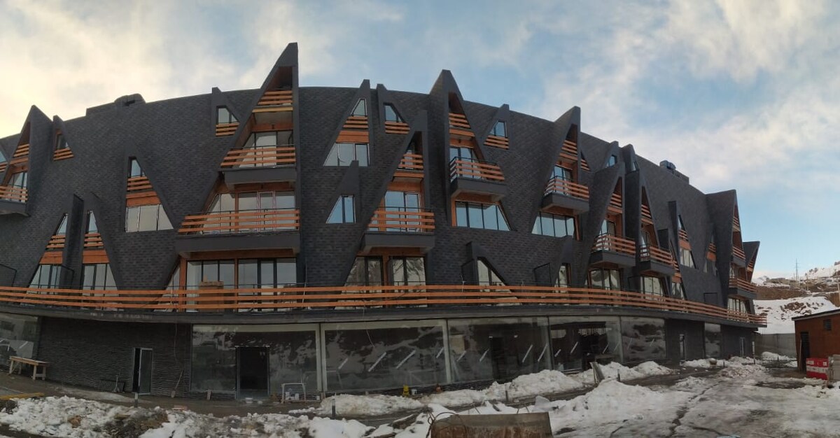 A modern building features a unique architectural design with a dark exterior and triangular windows. Balconies extend outward, providing outdoor access for the upper levels. The surrounding area is marked by patches of snow, indicating a winter environment.