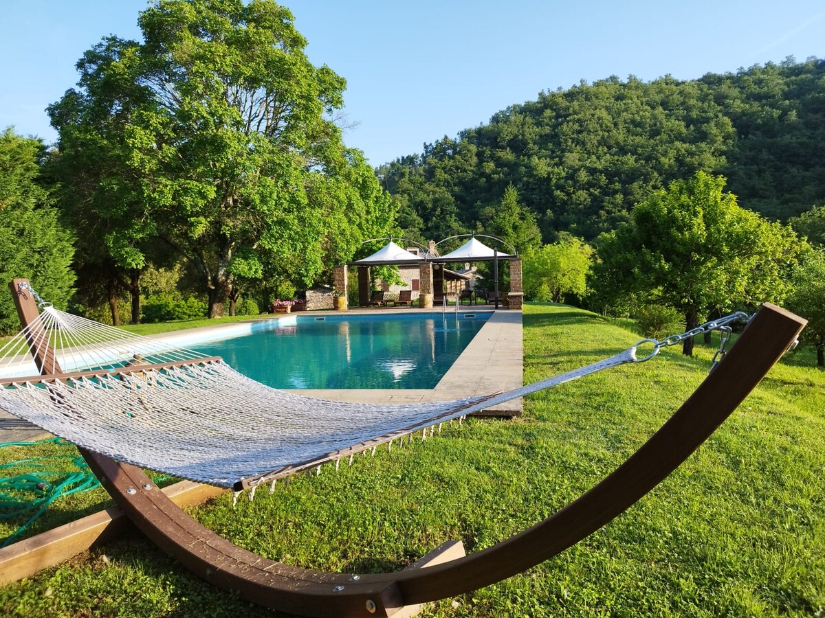 A hammock is positioned in a lush green area, gently curving beside a large swimming pool. Sunlight reflects off the water, highlighting the serene surroundings, which include trees and landscaped areas. A pavilion with white canopies is visible in the background, providing a shaded relaxation space.