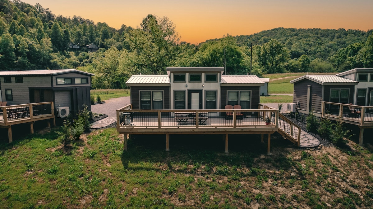 A row of tiny homes is displayed, each with a wooden deck surrounded by greenery. The sunset casts a warm glow over the scene, highlighting the natural landscape in the background and creating a serene atmosphere.