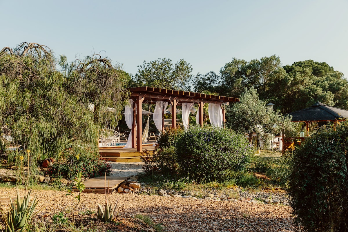 A serene outdoor area is shown, featuring a wooden pergola draped with light fabric. Surrounding greenery includes various trees and shrubs, with a stone path leading through a landscaped garden filled with natural textures and colors.