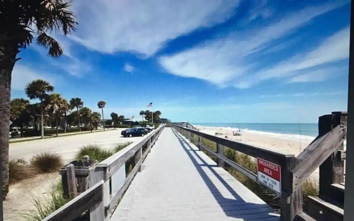 A wooden boardwalk stretches along the beachfront, bordered by palm trees and sea oats. The ocean is visible in the distance, with a clear blue sky overhead. A parked car and beach access sign can be seen along the path, indicating a welcoming coastal location.