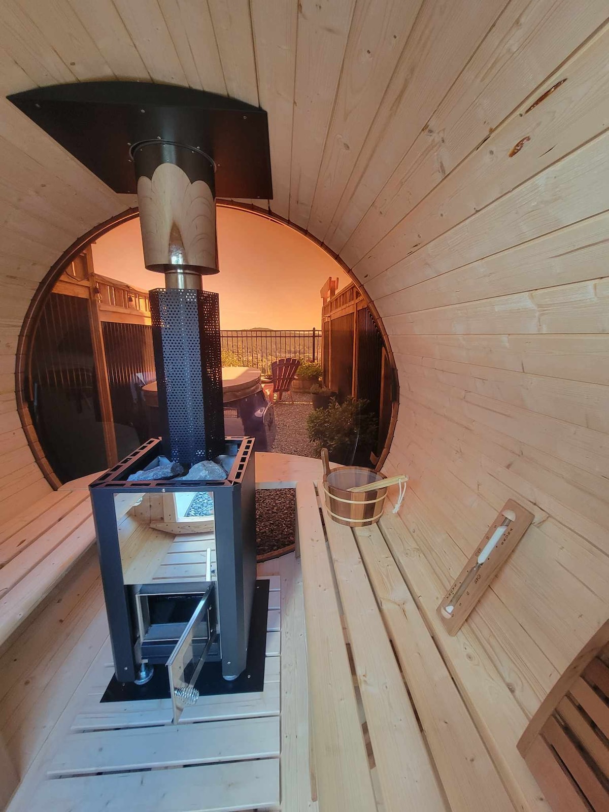 A barrel sauna interior is displayed, featuring wooden paneling and a central wood-burning stove. The warm glow from the sunset outside creates a calming atmosphere. Benches line the walls, providing comfortable seating for relaxation.