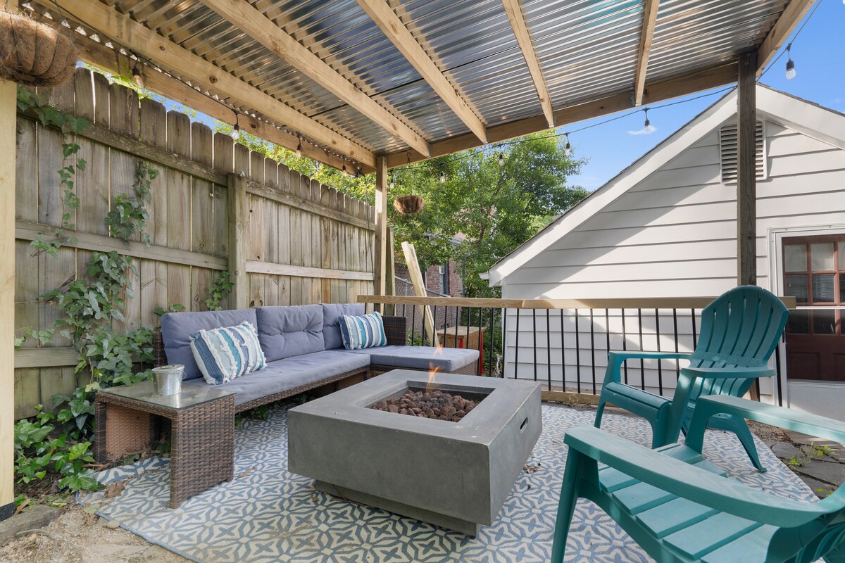 A covered patio area features comfortable seating with colorful cushions. A modern fire pit sits at the center, surrounded by a patterned rug. Greenery grows along the wooden fence, adding a touch of nature to the cozy outdoor space.