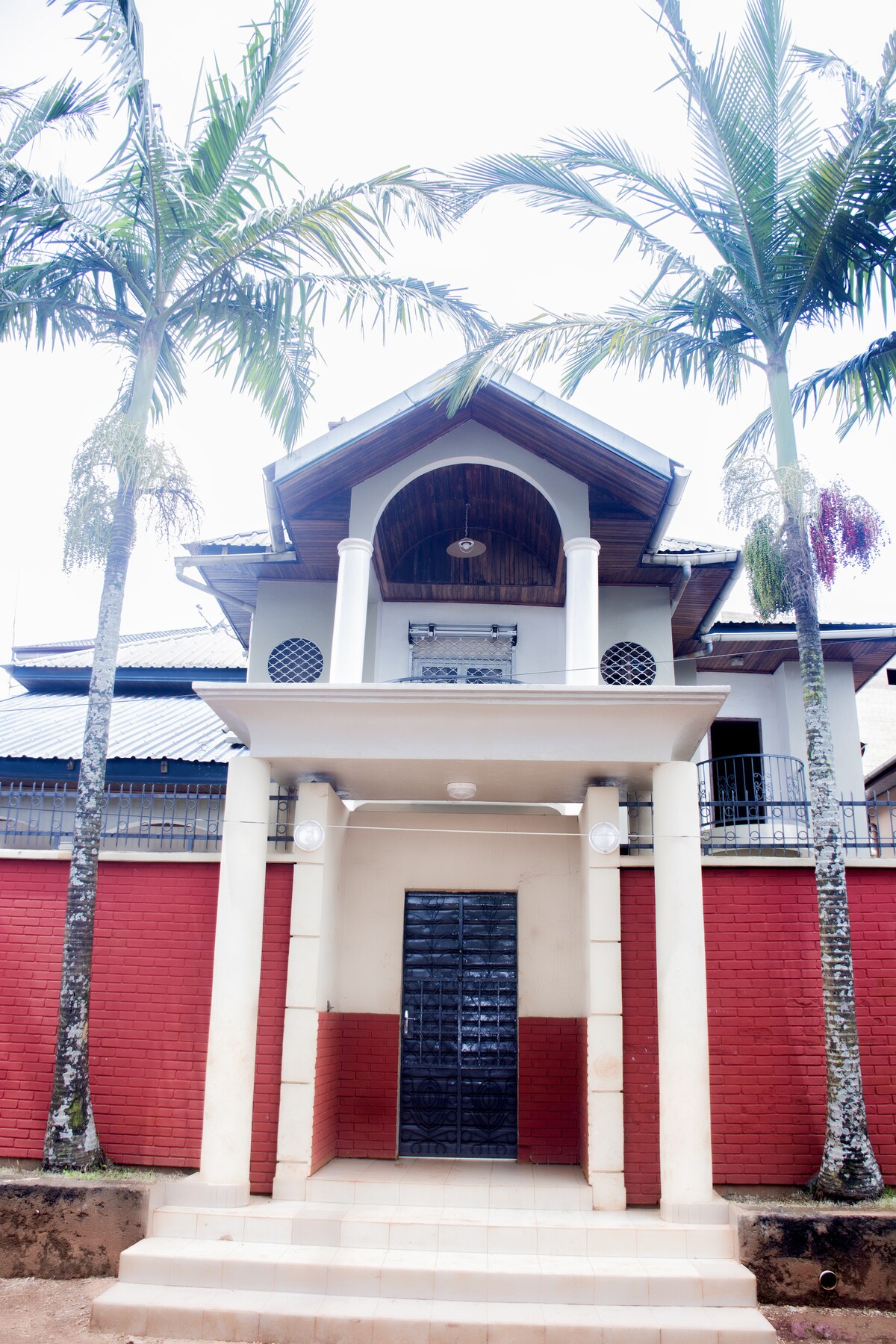 The exterior of the villa is showcased, featuring a symmetrical design with a central entrance door framed by elegant columns. Towering palm trees stand on either side, complementing the vibrant red brick walls. Above, a decorative overhang adds architectural interest to the facade.