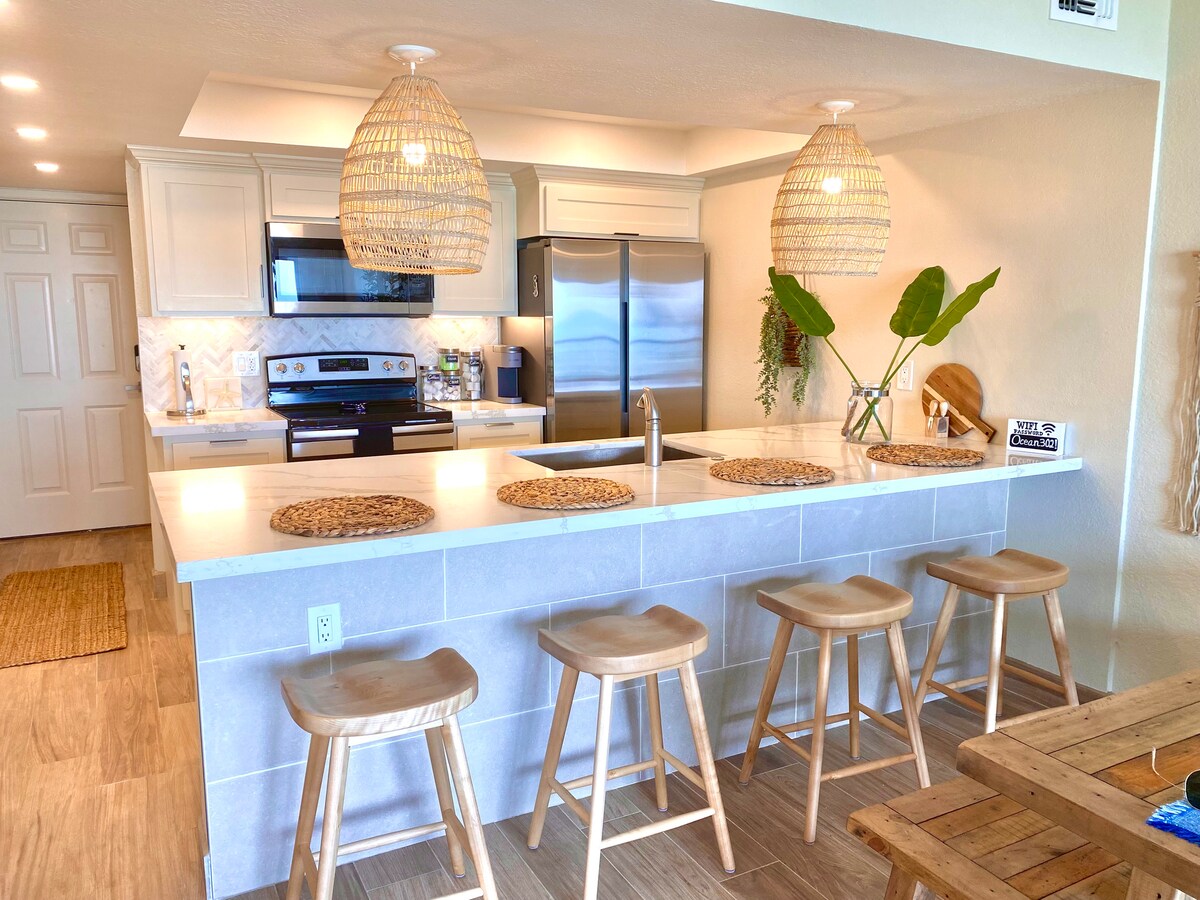 A modern kitchen displays an open layout with light cabinetry and stainless-steel appliances. Two unique pendant lights hang above a counter with four wooden stools. Natural elements are introduced by a green plant, enhancing the airy ambiance.