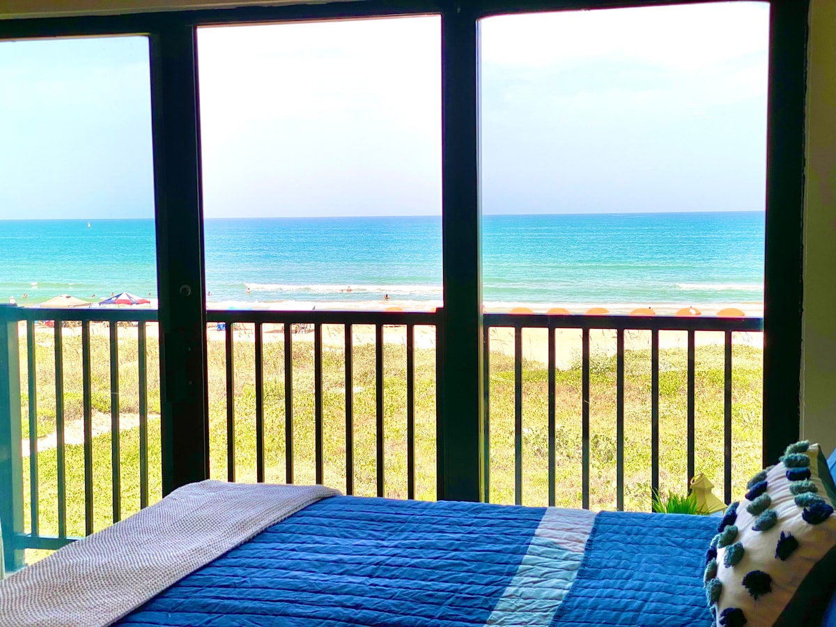 A spacious bedroom features a large window with views of the beach and ocean. Light turquoise waters contrast with golden sands, while beach umbrellas are visible in the distance. A blue quilt covers the bed, enhancing the coastal aesthetic.