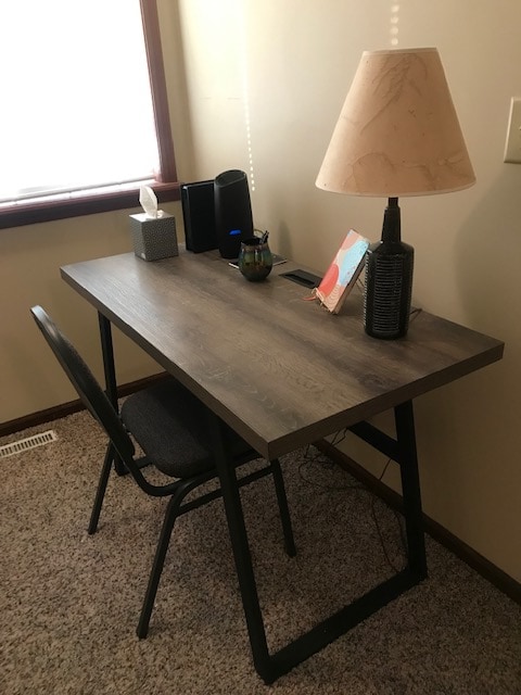 A simple workspace is featured with a sleek wooden desk and a matching chair. A lamp rests on the desk beside a small tissue box and a decorated picture. Natural light filters in through a nearby window, casting a soft glow across the area.