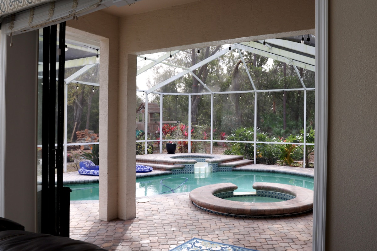 A screened-in pool area is visible, featuring a spacious pool with a built-in hot tub. Lush greenery and colorful plants surround the space, creating a serene environment. Natural light enters through the mesh enclosure, enhancing the inviting feel of the outdoor living area.