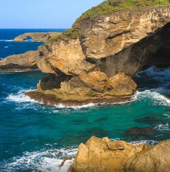 A rugged coastal landscape is depicted with striking rock formations jutting into the turquoise ocean. Waves crash against the rocks, creating a serene yet dynamic scene. The clear blue sky enhances the natural beauty of the coastline.