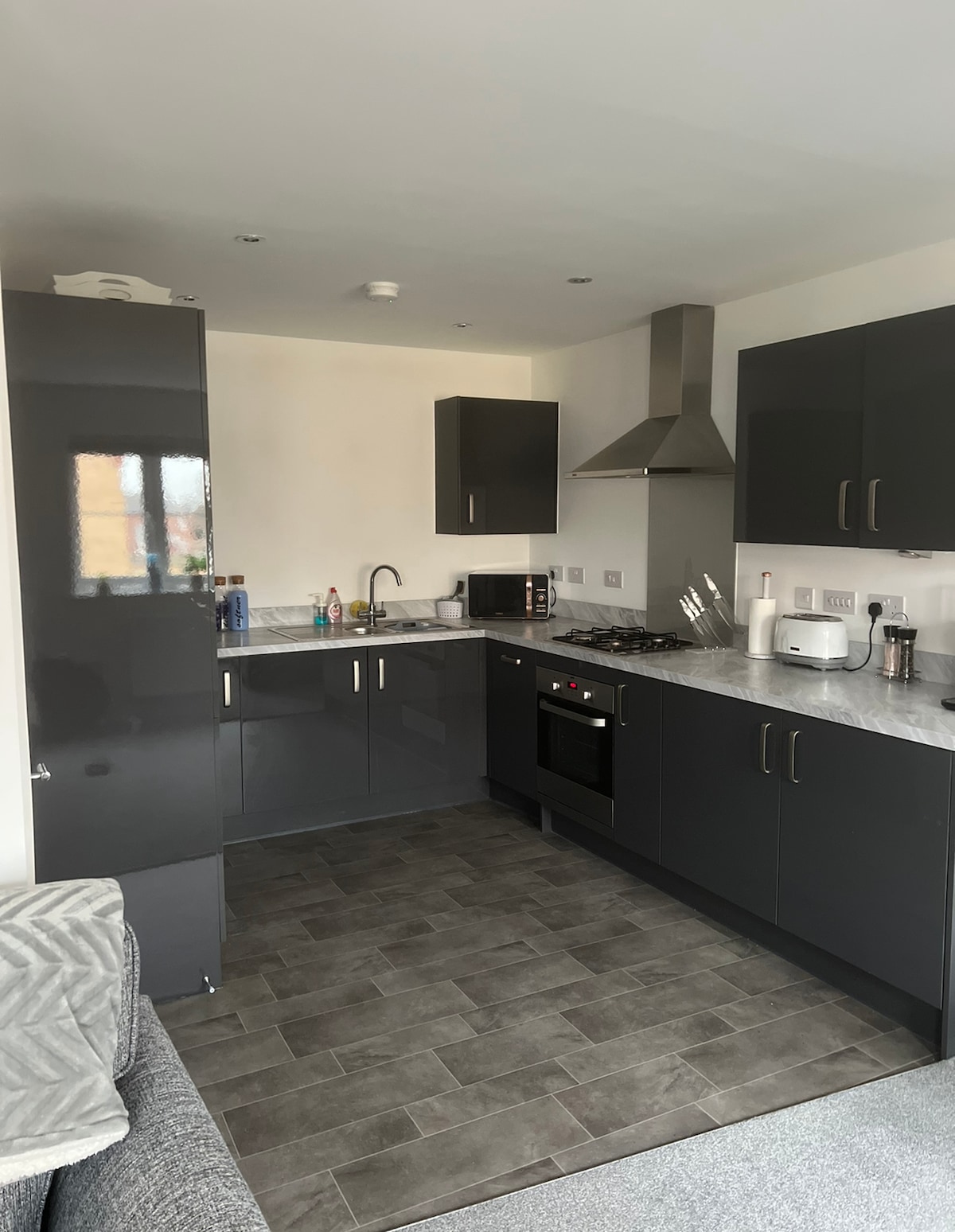 A modern kitchen featuring sleek black cabinetry and a marble countertop. Essential appliances are included, such as an oven, stovetop, microwave, and refrigerator. The space is well-lit with natural light coming through a window, providing a functional and inviting cooking area.