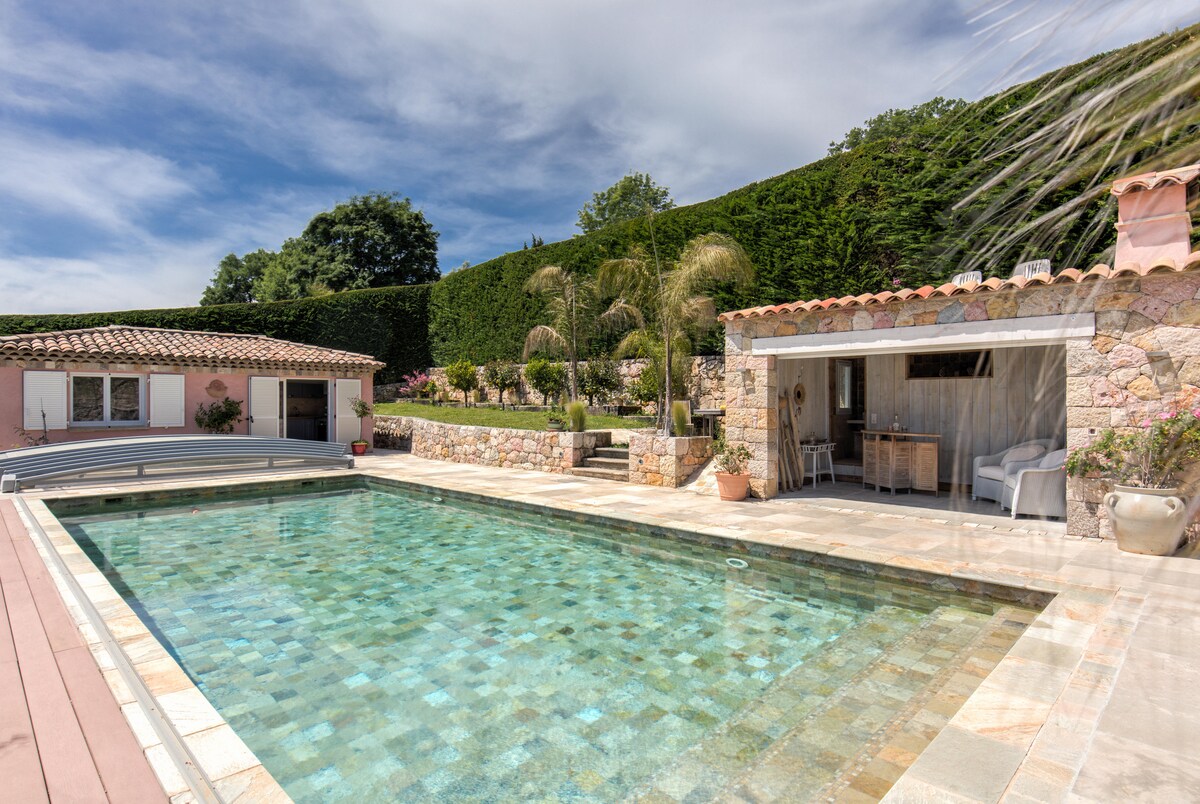 A spacious outdoor area features a large heated pool surrounded by a stone terrace. A pool house is visible to the side, providing an inviting space with seating. Lush greenery and landscaped gardens create a serene atmosphere, enhancing the overall enjoyment of the villa's exterior.
