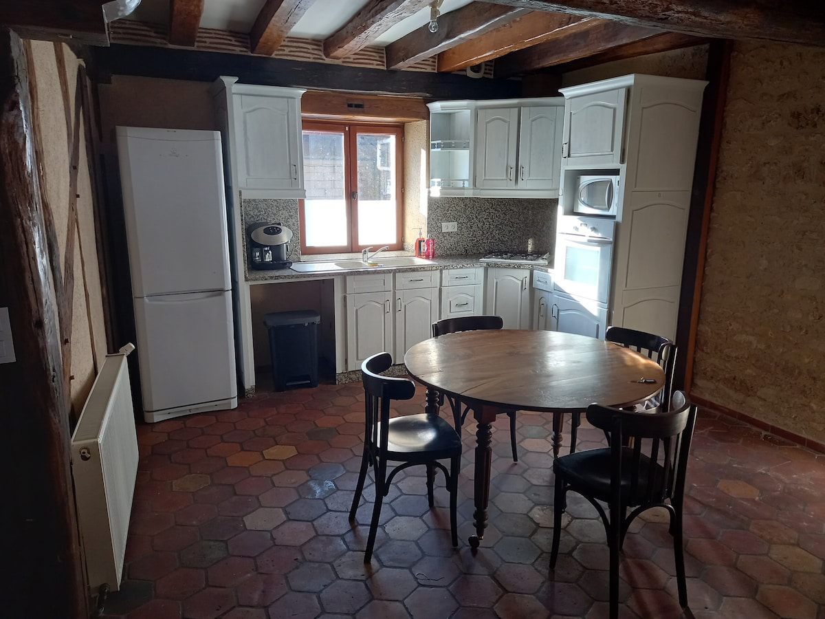 A well-equipped kitchen features a round wooden table surrounded by four black chairs. Cabinets and appliances, including a refrigerator and microwave, are visibly arranged along the walls. Natural light enters through a small window, illuminating the warm, rustic tile floor.
