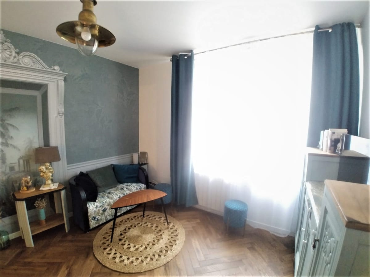A cozy living space is featured, showcasing a sofa adorned with floral patterns. A round wooden table sits in front, with a small blue stool nearby. Natural light fills the room through a large window, complemented by elegant curtains. Decorative elements include a rug and wall art.