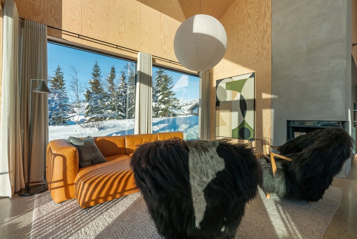 A spacious living area features large windows offering views of snow-covered trees. A modern orange sofa sits beside two black chairs with furry textures. A graphic art piece is visible on the wall, alongside a soft area rug on the concrete floor.