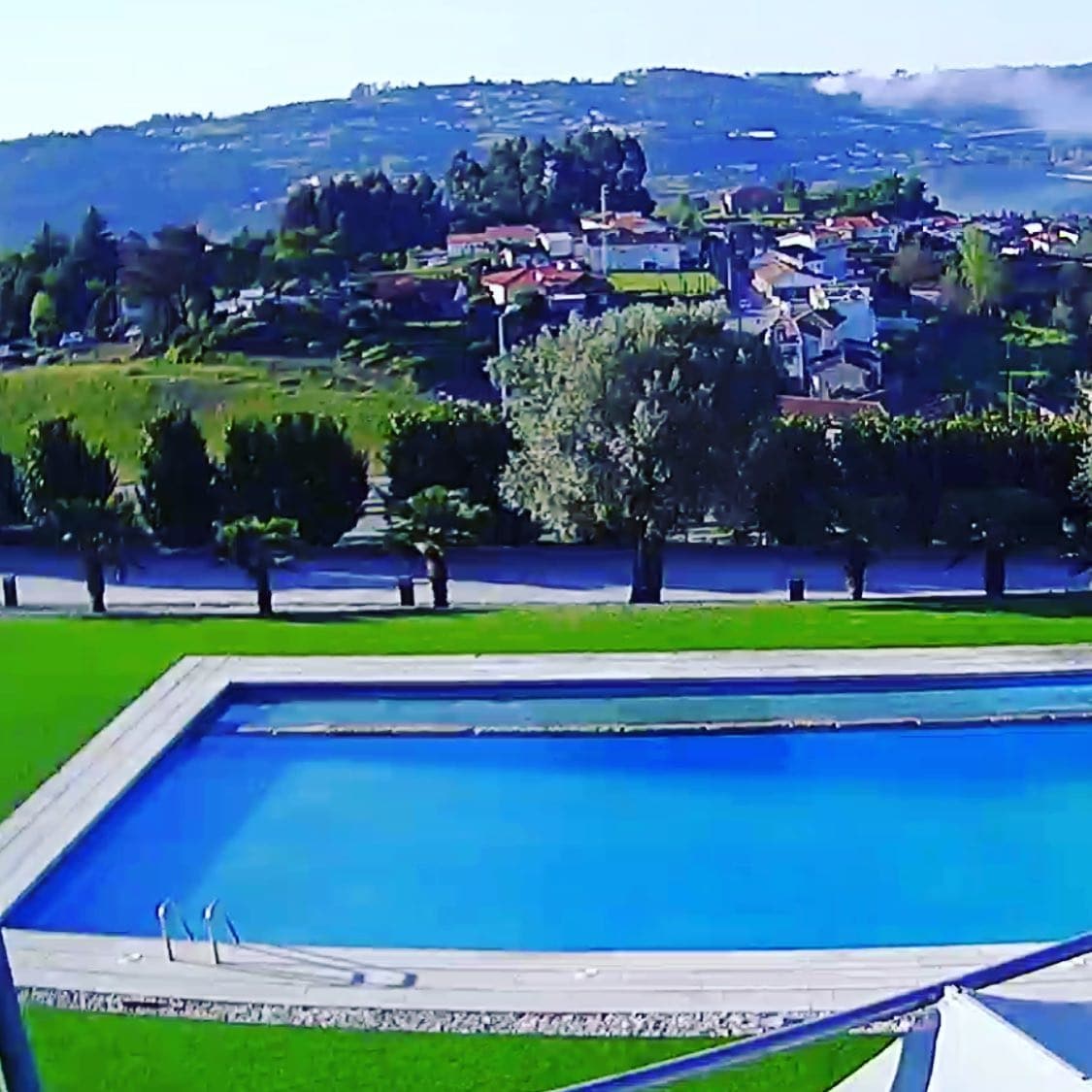 A tranquil outdoor space features a large swimming pool surrounded by meticulously maintained grass. In the background, hills and quaint homes create a serene landscape under a clear sky.