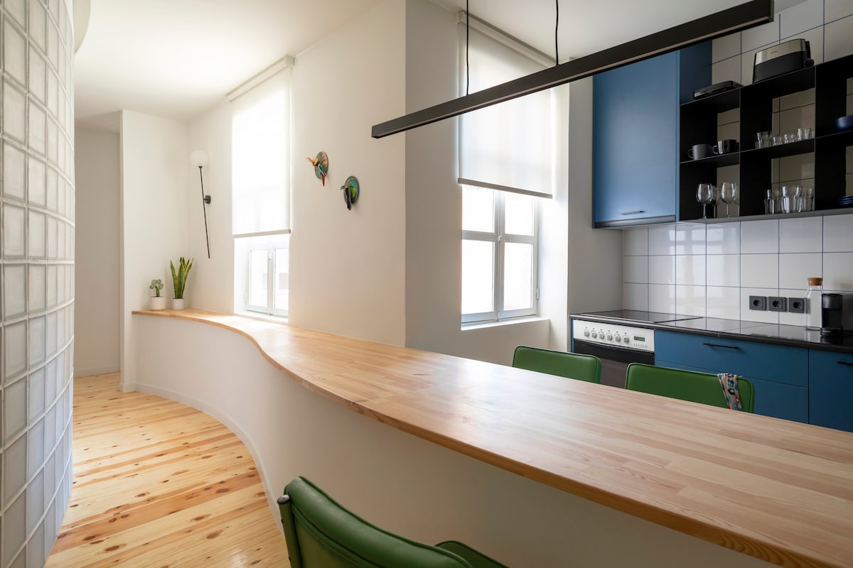 A modern kitchen features a curved wooden countertop with several green chairs arranged at it. Natural light is provided by two large windows with softly diffused shades. The kitchen cabinetry includes blue upper cabinets and white lower tiles, complementing the sleek appliances.