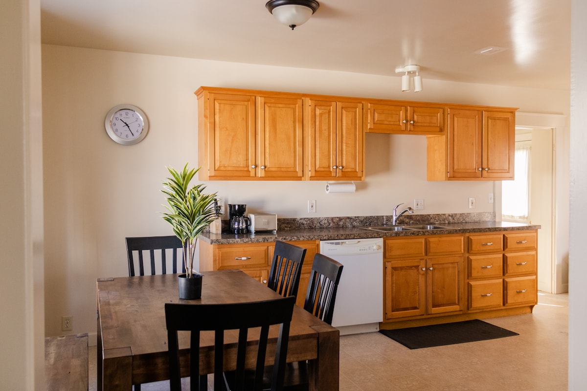 A bright kitchen area features wooden cabinetry and a granite countertop, complemented by a dining table with black chairs. A clock and a small plant add to the decor. Appliances include a dishwasher and a coffee maker, providing convenience for meal preparation.