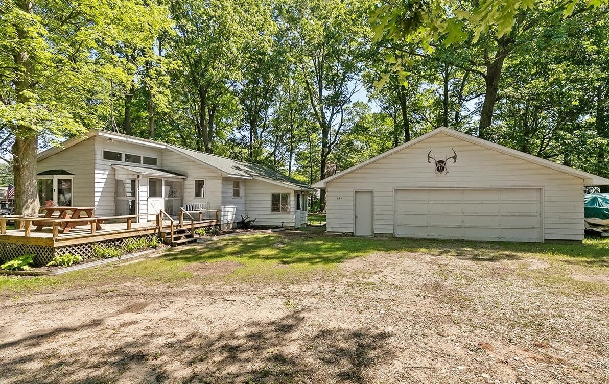 A spacious, newly renovated cottage is visible, showcasing a large deck at the entrance. Adjacent, a garage with a decorative motif is present. The surroundings are framed by trees, contributing to a serene outdoor environment. The gravel driveway provides ample parking.