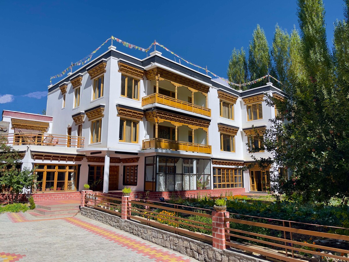 A three-story building features traditional architectural details, including wooden balconies and intricate carvings. Surrounding garden beds are filled with greenery and flowers. The foreground presents a paved pathway, leading to the entrance, under a clear blue sky.