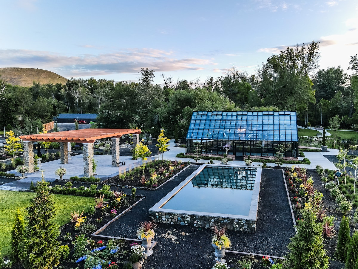 An aerial view captures the Gardens at The Brick House Gardens & Nursery, featuring a glass conservatory, rectangular reflection pool, and a stone pergola. Lush landscaping surrounds the pathways, enhancing the serene setting, with well-maintained plants and trees adding to the tranquil atmosphere.