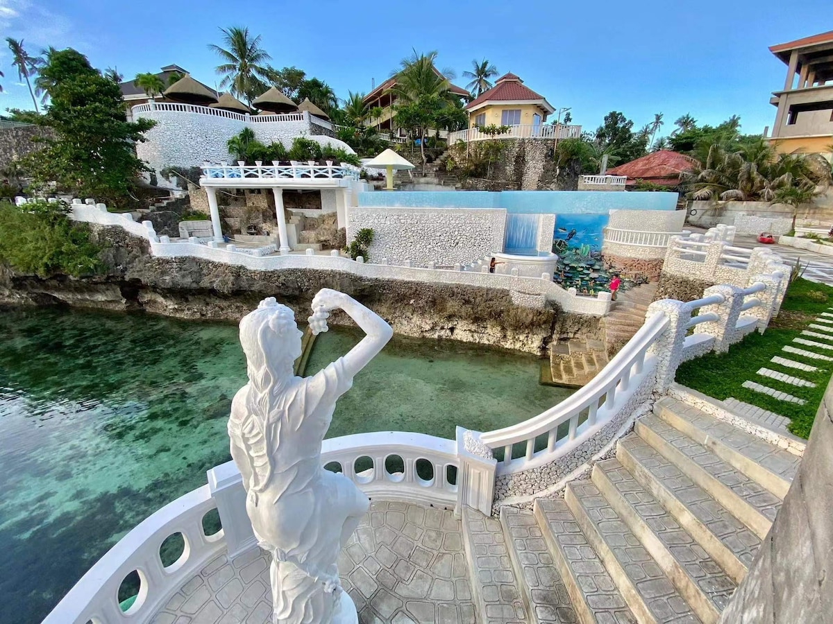 A statue stands elegantly at the edge of a stone staircase leading down to a tranquil oceanfront area. Lush greenery surrounds the scene, while a swimming pool is visible in the background, complemented by well-maintained walkways and cliffside architecture.