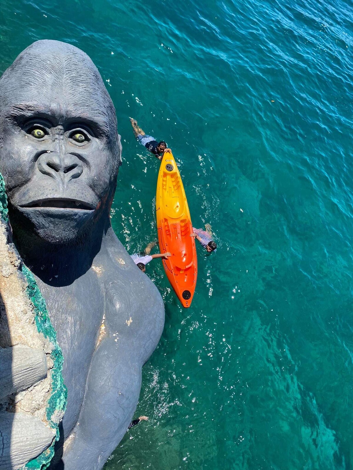 An overhead view captures individuals interacting with a bright orange kayak in clear, blue water. The kayak is positioned near a rocky surface, with waves gently lapping against the sides. Nearby figures are visible, enjoying the outdoor experience in a vibrant coastal setting.