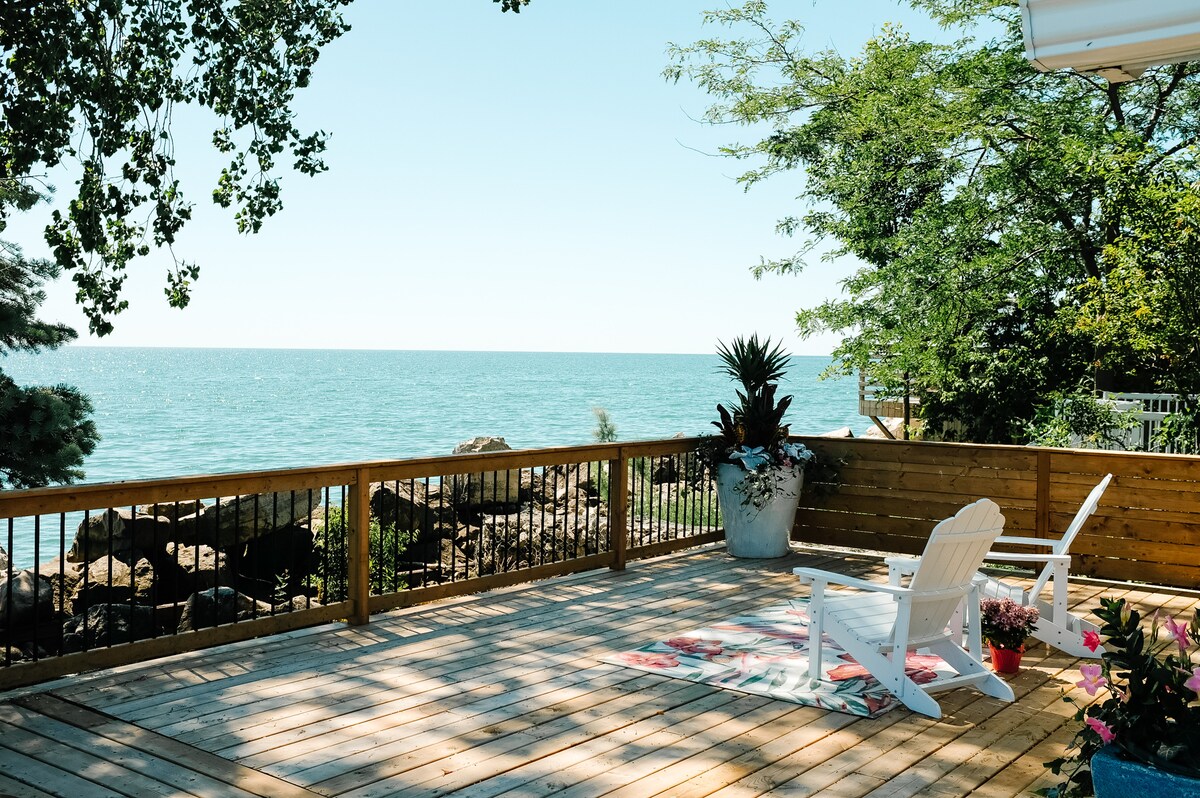 A spacious wrap-around deck offers unobstructed views of Lake Erie. Two white Adirondack chairs are positioned on the deck, surrounded by lush greenery and potted plants. The sun casts a gentle light on the wooden flooring, enhancing the serene waterfront setting.