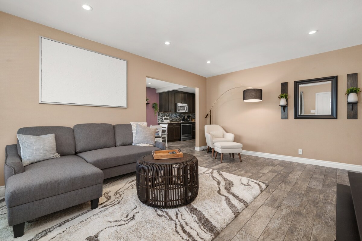A comfortable living area is furnished with a gray sectional sofa and a circular coffee table. A cozy reading chair is positioned nearby, with soft lighting provided by a modern lamp. A neutral color palette complements the textured area rug and the warm wall tones.