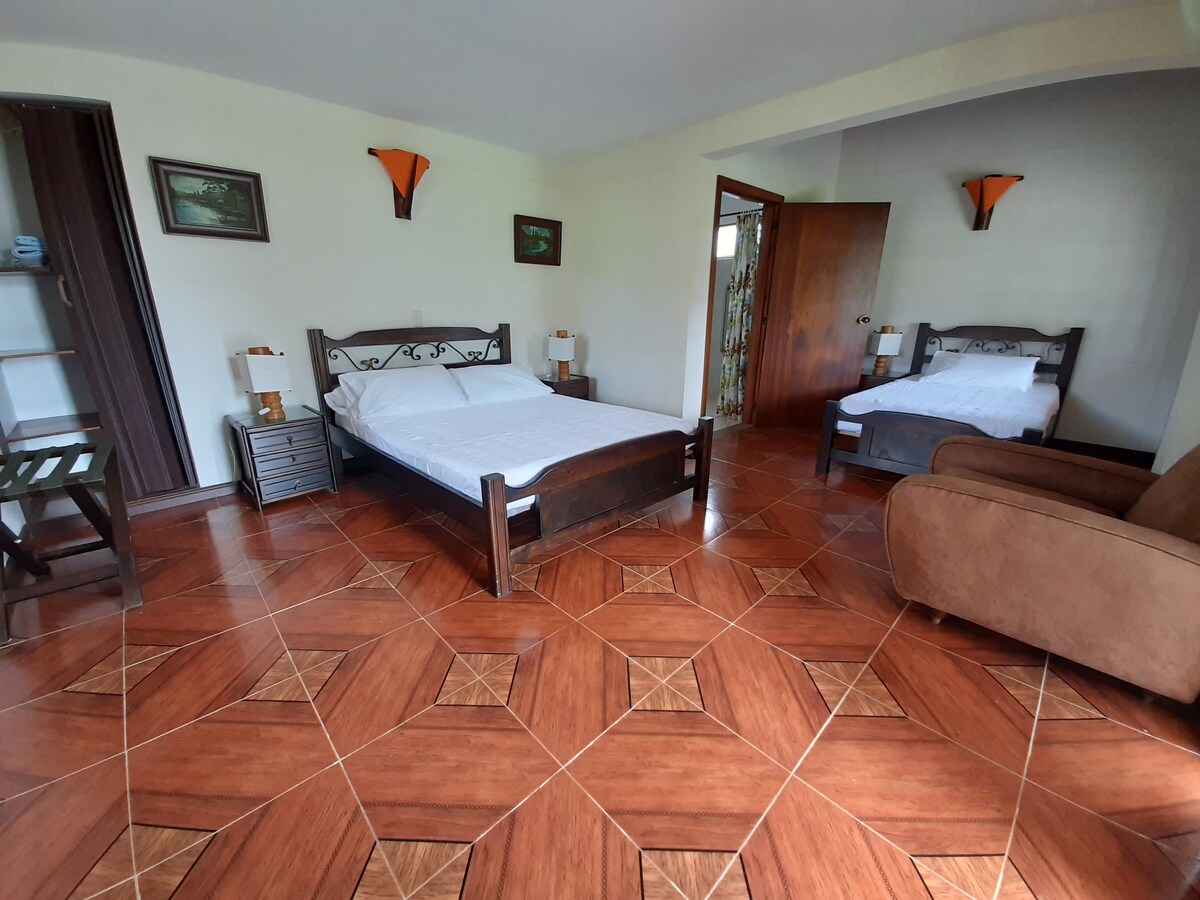 A spacious room is depicted, featuring a large bed and a smaller bed, complemented by a brown sofa. Light wood furniture, including a nightstand and a small chair, enhances the interior. Natural light filters through the doorway, illuminating the tiled floor.