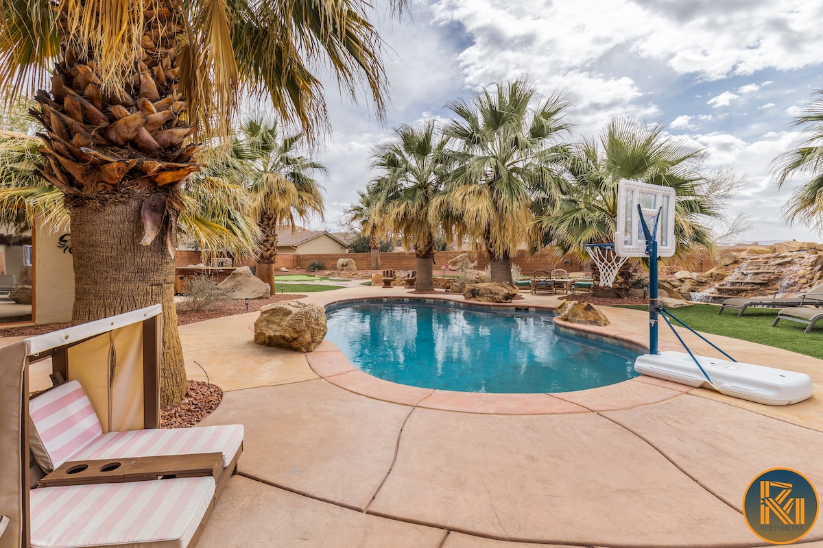 A vibrant outdoor area features a curved saltwater pool surrounded by lush palm trees. A basketball hoop stands beside the pool, with rock formations visible in the background. Comfortable lounge chairs are positioned nearby on the patio, inviting relaxation.