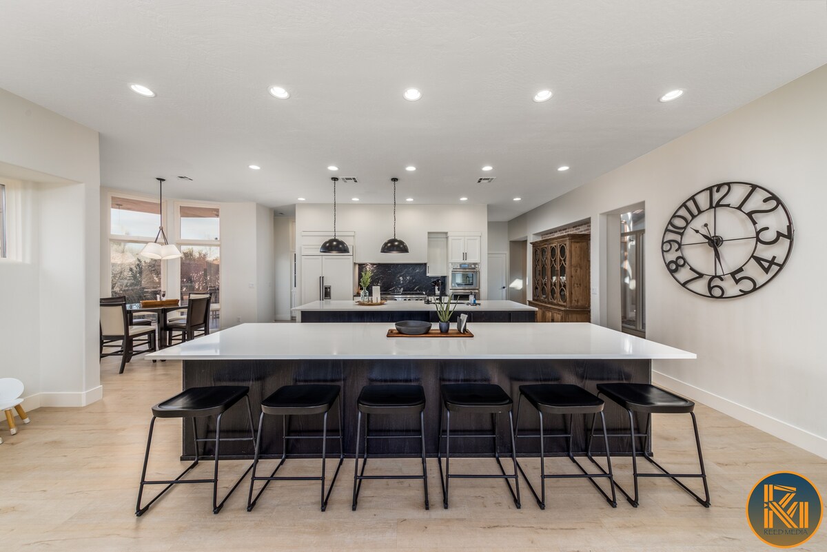 A modern kitchen features a large central island with seating for six. Sleek black bar stools are positioned around the island. An elegant clock adorns the wall, while natural light fills the space through large windows, highlighting the clean lines and contemporary design.