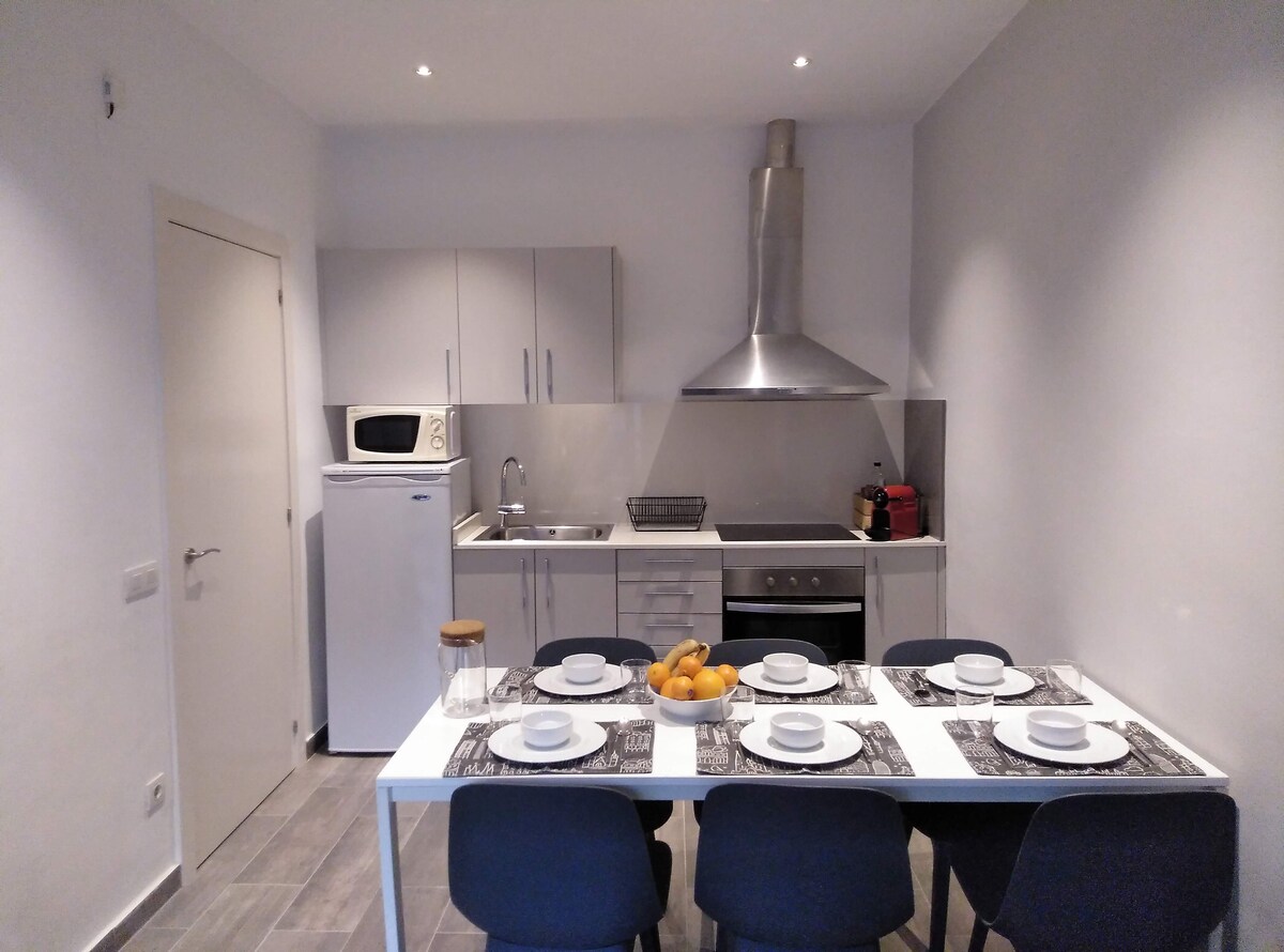 A modern kitchen with sleek cabinetry and stainless steel appliances is shown. A dining table set for six is displayed, featuring a neutral table runner and a bowl of oranges. Ample natural light is present, enhancing the space's functionality.