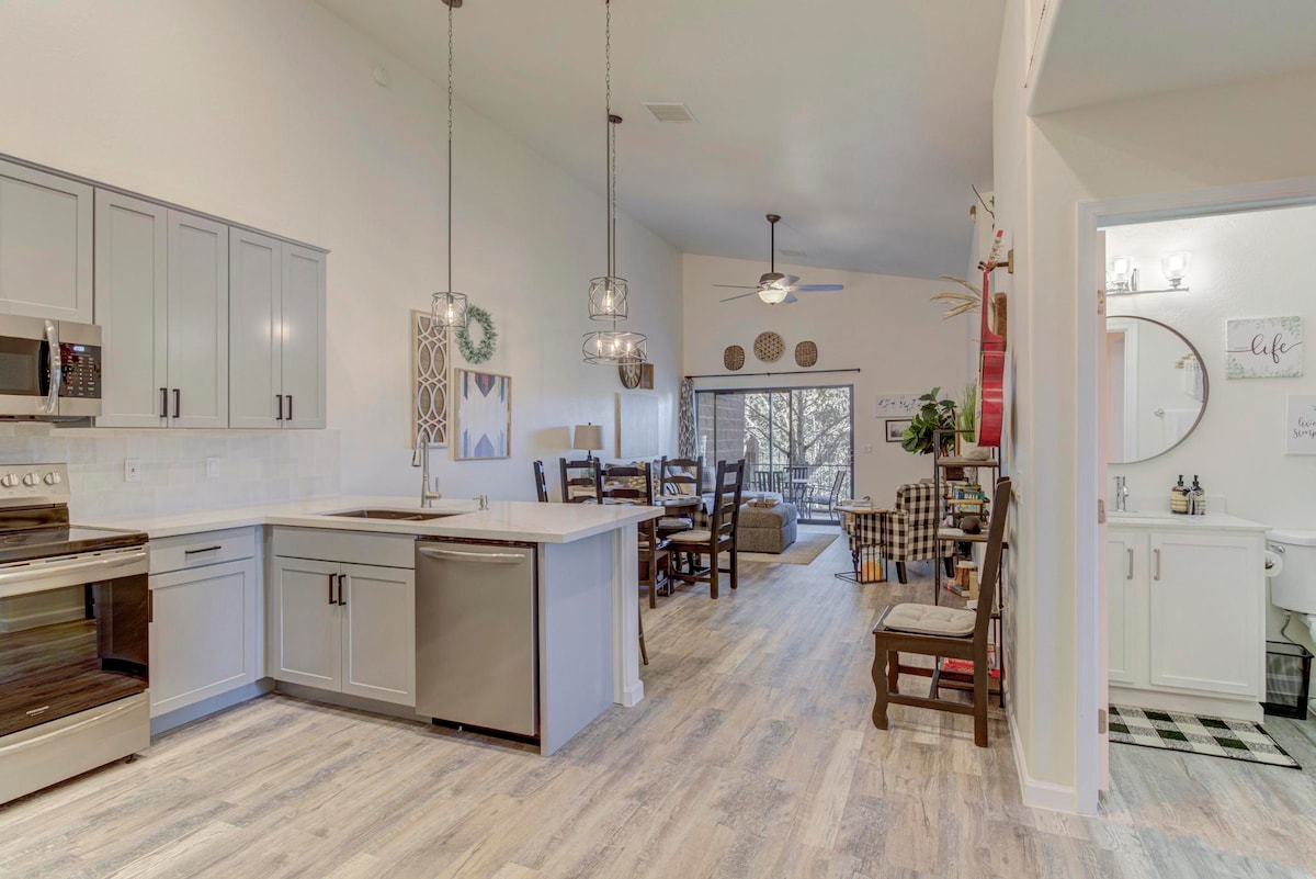 The open kitchen features modern appliances, including a stainless steel stove and dishwasher, surrounded by light cabinetry and white countertops. A spacious dining area is visible, with a large table and wooden chairs. Natural light fills the space, creating a welcoming atmosphere.