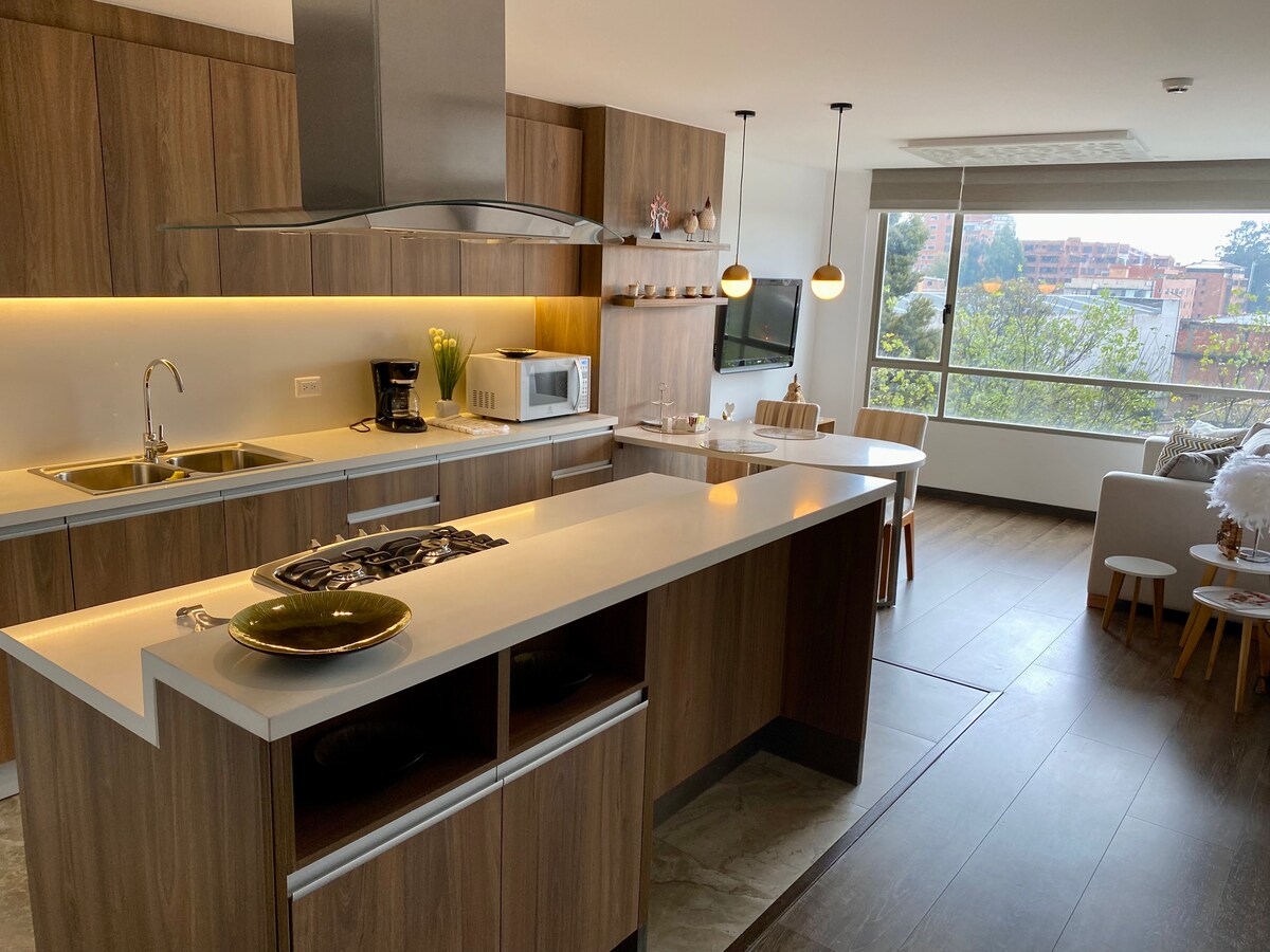 The modern kitchen features wood cabinetry and a spacious island, equipped with a gas stove, sink, and microwave. Natural light fills the area from large windows, complemented by pendant lighting. A small dining table with wooden stools is visible in the background, enhancing the open layout.