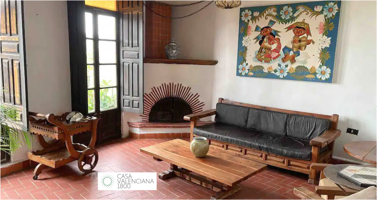 A welcoming living area features a dark wooden sofa and a natural wood coffee table. A colorful wall art piece depicting two figures is displayed above a decorative fireplace. Wooden doors provide a connection to the outdoor space, enhancing the room's natural light.