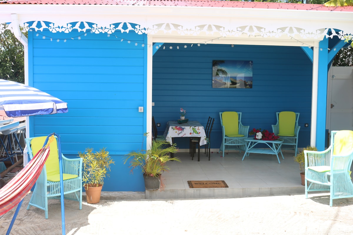 An inviting outdoor seating area features bright blue walls with white accents. Two colorful chairs and a small table create a cozy space beneath a covered patio. Decorative elements, including potted plants and a hammock, enhance the relaxing atmosphere.