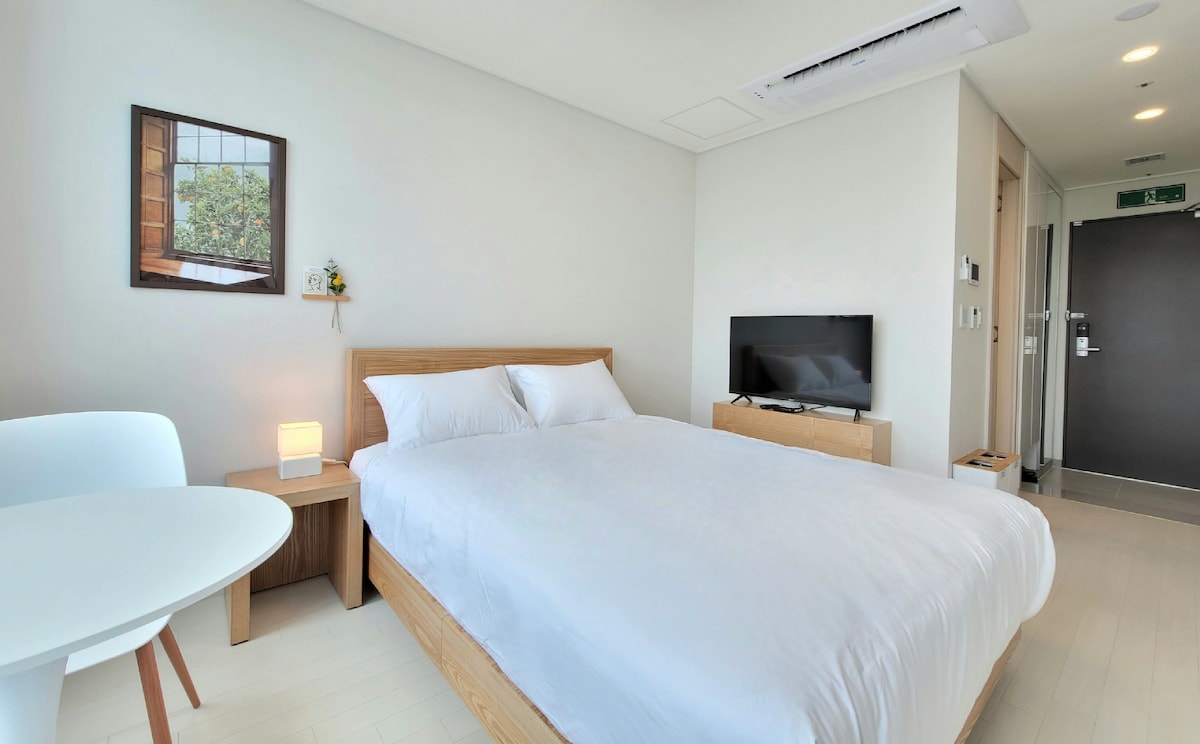 A modern room is presented with a queen-sized bed covered in pristine white bedding. A small wooden table for two is positioned beside the bed. A flat-screen TV is mounted on the wall opposite the bed, and a simple artwork adorns the wall above.