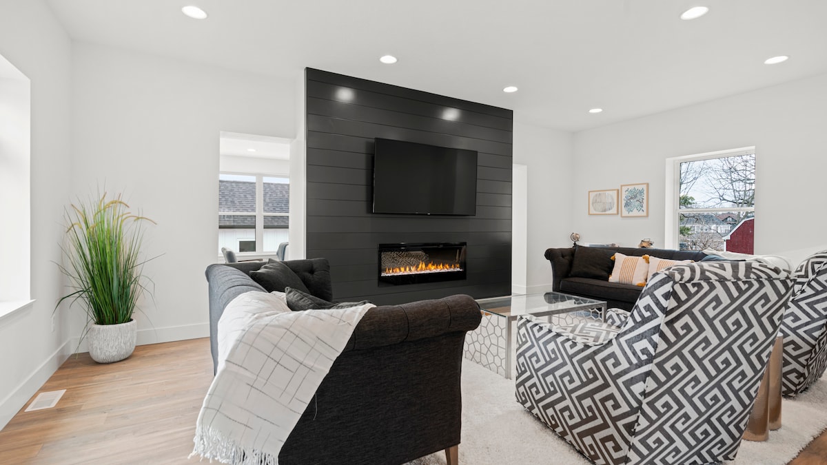 A spacious living area features a modern black accent wall housing a flat-screen TV above an electric fireplace. Comfortable seating is provided by a dark sofa and patterned armchairs. Natural light enters through multiple windows, enhancing the inviting space decorated with a potted plant.
