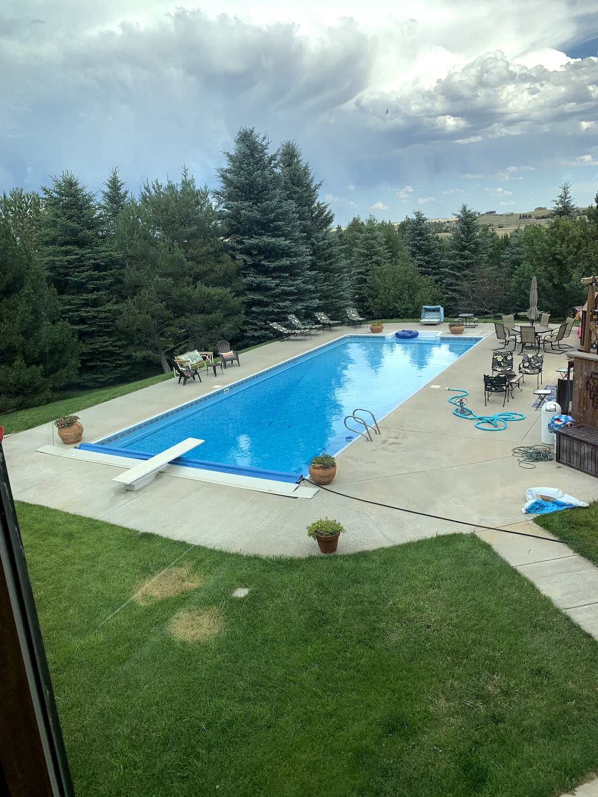 A spacious outdoor pool area is surrounded by tall green trees, creating a private atmosphere. Loungers are arranged along the poolside, and a dining area with tables and chairs is visible in the background. Pool floats are seen floating in the clear water.