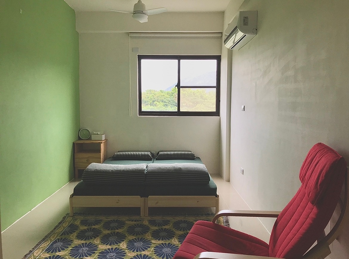A bright and airy room is presented with a large window offering views of the surrounding hills. A red chair complements a simple wooden bed, which can be separated into twin beds. A patterned rug adds a touch of color to the light-toned space.