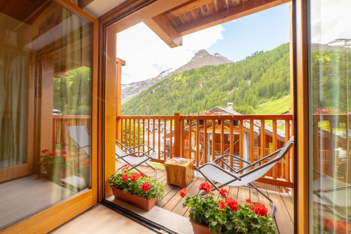 A balcony is depicted with two lounge chairs positioned for relaxation, overlooking a mountain landscape. Bright red flower pots adorn the balcony, complementing the wooden railing. The mountains in the background are partially covered with greenery, creating a peaceful outdoor space.