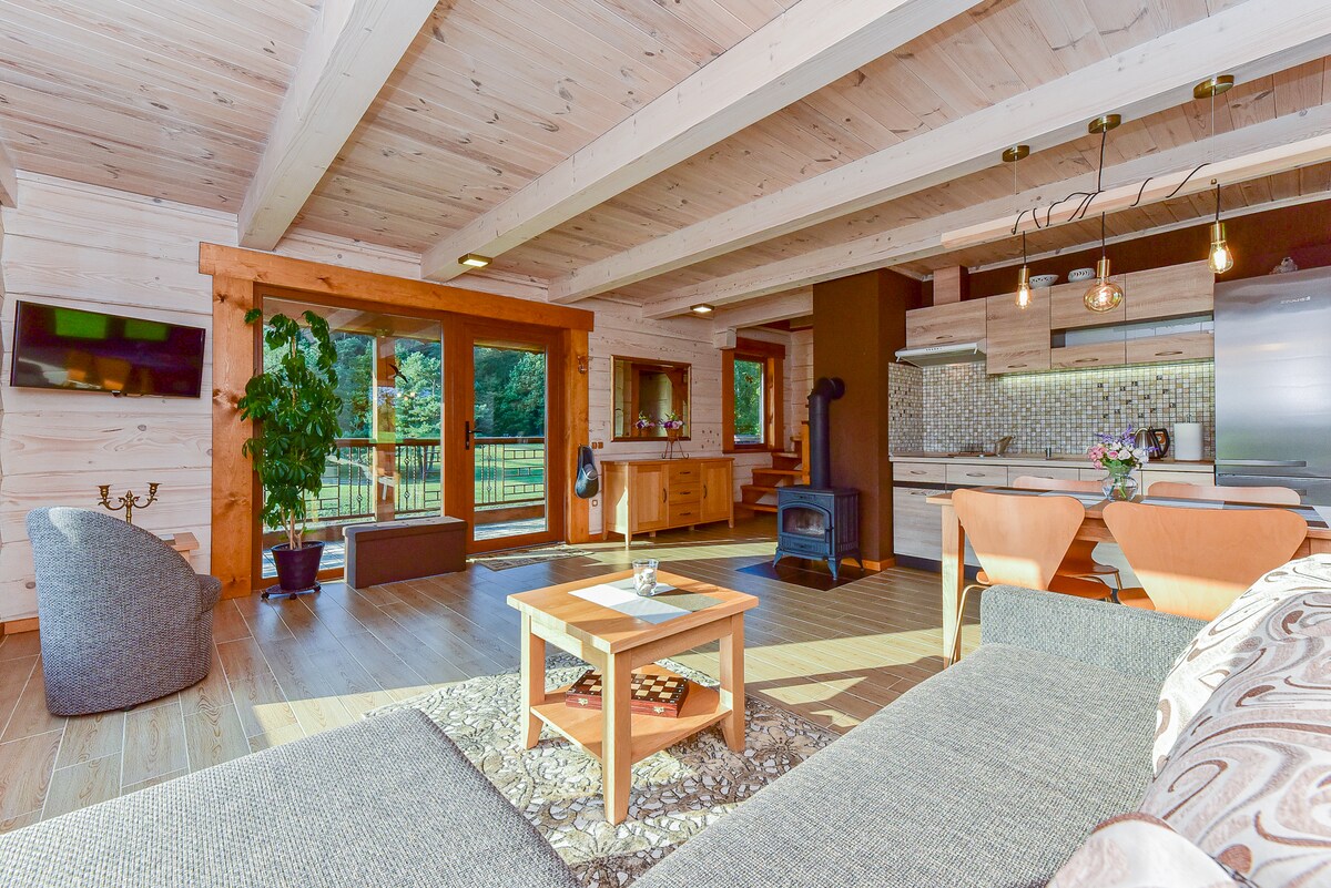 An open living area features wooden beams and large glass doors that provide a view of the outdoor space. A comfortable sofa is positioned near a small coffee table, while a stove is situated in a corner. The kitchen area is equipped with modern cabinetry and appliances.